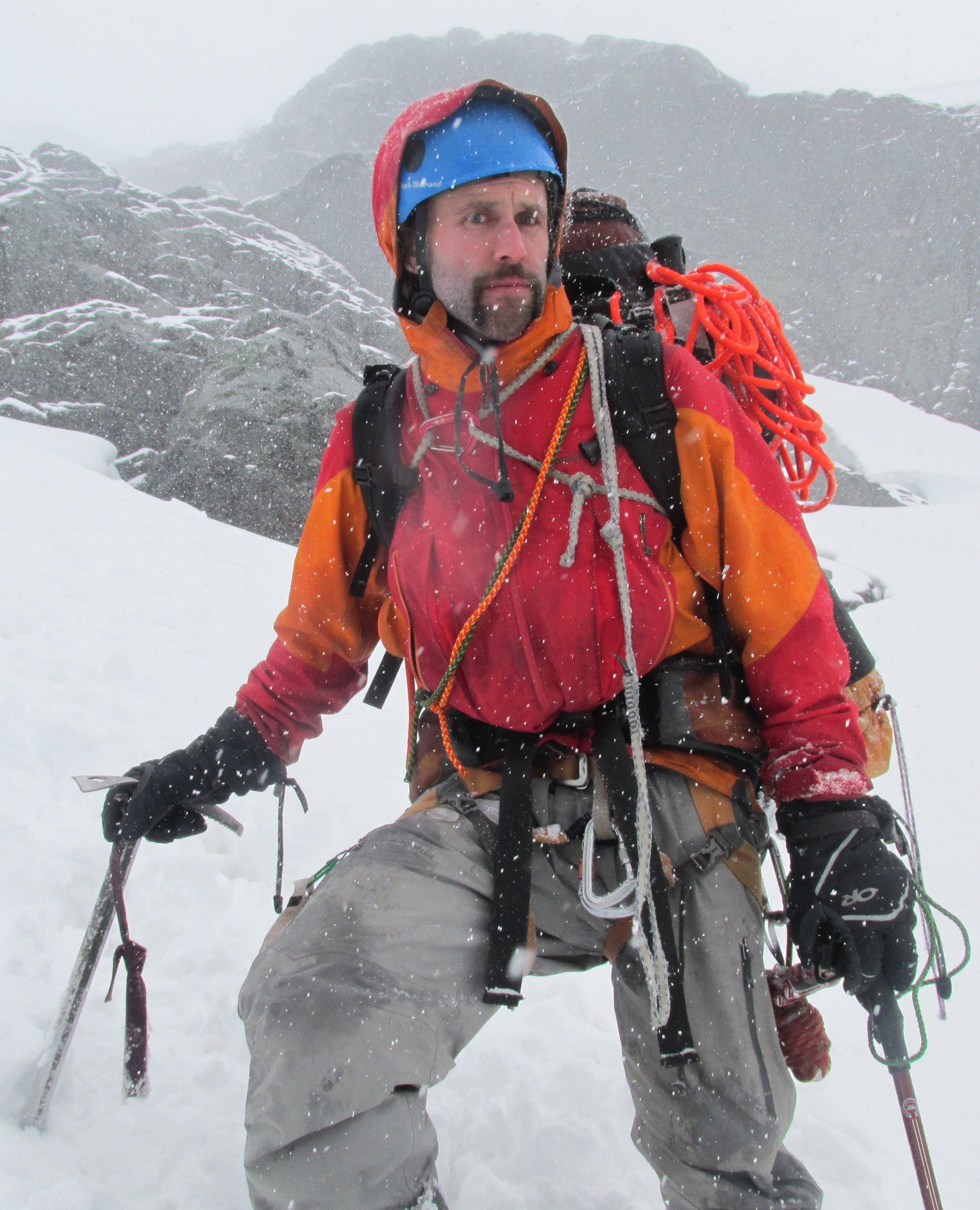 jared on a snow slope in North Cascades National Park. Helmet, ice axe, loaded pack, rope, snow, overcast skies