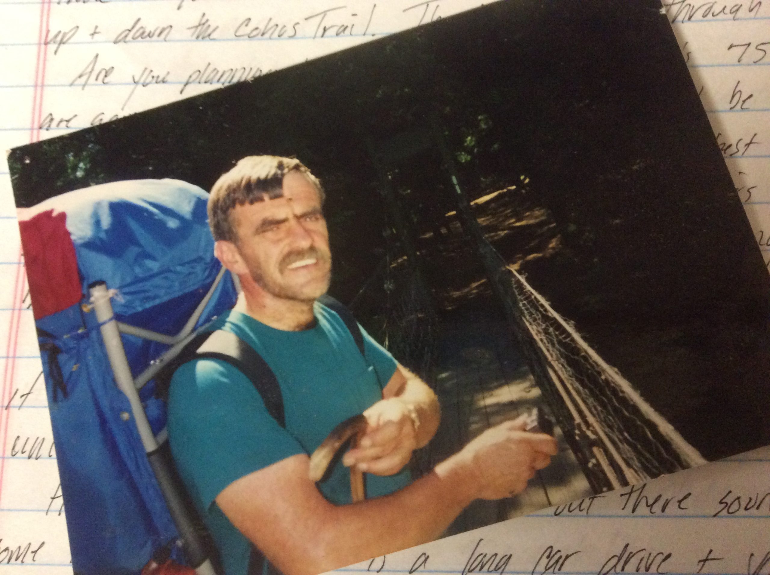 a white male with a mustache and brown hair.  He is wearing a large blue backpack and standing on a narrow footbridge.  He is smiling